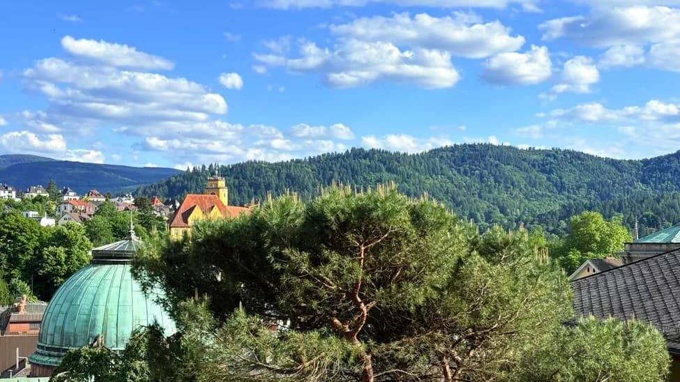 Vue de Baden-Baden et des collines boisées de la Forêt-Noire sous un ciel bleu, avec le dôme vert d'un bâtiment thermal au premier plan